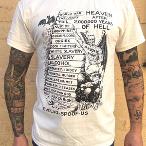 Può includere: T-shirt color crema con un design grafico nero. Il design include una figura di diavolo, un elenco di mali sociali e il testo "Heaven After 2,000,000 Years of Hell". La maglietta presenta anche il testo "Evolvo-Spoof-Us".