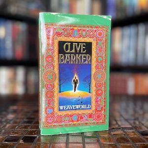 May include: A hardcover book titled "Weaveworld" by Clive Barker. The cover features a person walking towards a bright light, with the title and author's name in gold lettering. The book has a green border with colorful circular designs.