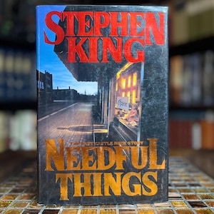 Könnte beinhalten: Ein gebundenes Buch mit dem Titel Needful Things von Stephen King. Das Cover zeigt eine dunkle Straßenszene mit einem Schaufenster, in dem ein Open-Schild und eine Puppe zu sehen sind. Der Autorenname ist in großen roten Buchstaben gedruckt.