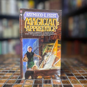 Pode incluir: Um livro de bolso intitulado "Magician Apprentice" de Raymond E. Feist. A capa apresenta uma ilustração de fantasia de um mago e um jovem. O livro inclui uma citação do The Washington Post Book World.