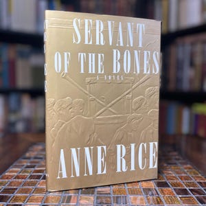 Peut inclure: Une couverture de livre dorée avec le titre "Servant of the Bones" et le nom de l'auteur "Anne Rice" en texte blanc. La couverture présente une image en relief de personnes portant une croix.