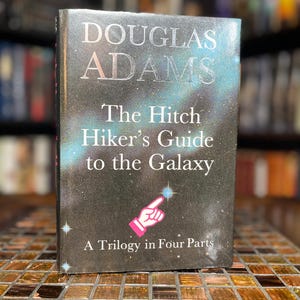May include: A hardcover book titled The Hitchhiker's Guide to the Galaxy by Douglas Adams, with the subtitle A Trilogy in Four Parts. The black cover features a cosmic nebula design with white stars and a pink pointing finger icon graphic.