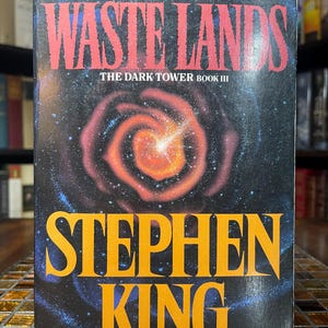 May include: A book cover for "The Waste Lands" by Stephen King, part of "The Dark Tower Book III." The cover features a cosmic design with a swirling orange and red galaxy against a dark blue background. The title and author's name are in bold, contrasting colors.