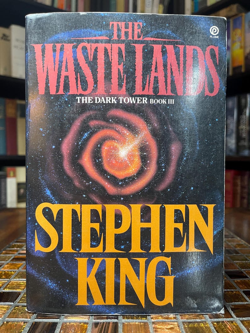May include: A book cover for "The Waste Lands" by Stephen King, part of "The Dark Tower Book III." The cover features a cosmic design with a swirling orange and red galaxy against a dark blue background. The title and author's name are in bold, contrasting colors.