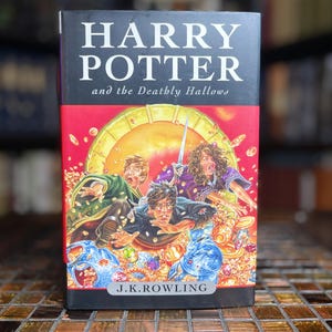 May include: A hardback book titled Harry Potter and the Deathly Hallows by J.K. Rowling. The cover features a vibrant illustration of three characters surrounded by gold coins and treasures against a bright yellow and red background.