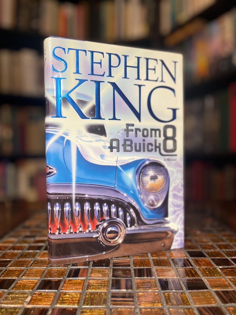 Może przedstawiać: Twarda oprawa książki "From a Buick 8" Stephena Kinga. Okładka przedstawia niebieski klasyczny samoch&oacute;d z chromowanymi detalami na jasnym tle. Książka leży na powierzchni z mozaiki.