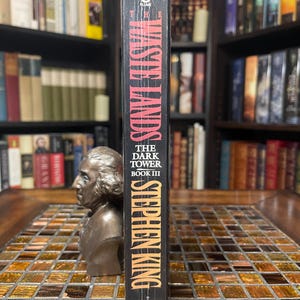May include: A hardcover book titled "The Waste Lands" by Stephen King, part of "The Dark Tower Book III" series. The spine is black with orange and white lettering. The book is standing upright on a mosaic tile surface.