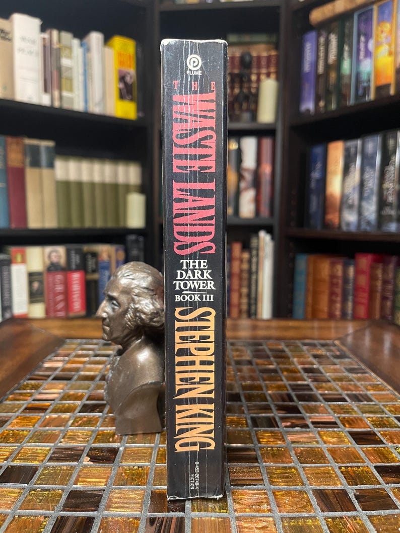 May include: A hardcover book titled "The Waste Lands" by Stephen King, part of "The Dark Tower Book III" series. The spine is black with orange and white lettering. The book is standing upright on a mosaic tile surface.