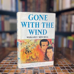 May include: A vintage paperback copy of the novel "Gone With The Wind" by Margaret Mitchell. The cover features a colorful illustration of a couple in a horse-drawn carriage with a background of a bustling city street. The text "Greatest best-seller of our century - the modern American classic already read by over 20,000,000 people." is printed at the bottom of the cover.