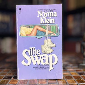 Peut inclure: Un livre de poche intitulé The Swap de Norma Klein avec une couverture violet clair. La couverture présente une illustration d'une personne allongée dans le cadre d'une fenêtre de voiture et une paire de chaussures de bébé blanches.