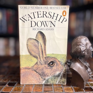 May include: A paperback book titled Watership Down by Richard Adams, featuring a brown rabbit illustration on the cover, the text World Number One Bestseller, and a small orange penguin logo, positioned next to a bronze-colored bust of a person.