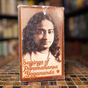 Może przedstawiać: Zabytkowa książka zatytułowana "Sayings of Paramahansa Yogananda" z portretem autora. Książka ma brązową okładkę z pomarańczowymi literami i kwiatowym wzorem. Książka leży na powierzchni z płytek mozaikowych.