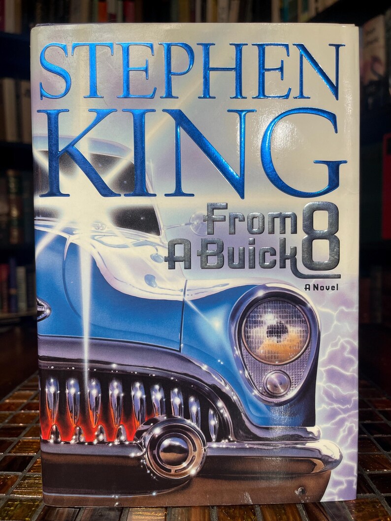 Może przedstawiać: Okładka książki "From A Buick 8" Stephena Kinga. Przedstawia niebieski klasyczny samoch&oacute;d z chromowanymi detalami i błyskawicą. Imię autora jest w dużych, błyszczących, niebieskich literach.