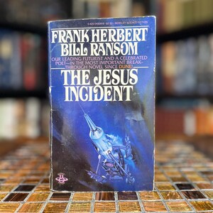 Puede incluir: Un libro de bolsillo titulado The Jesus Incident de Frank Herbert y Bill Ransom. La portada azul oscuro presenta el título y los nombres de los autores en blanco, junto a una ilustración central de un cohete plateado en el espacio.