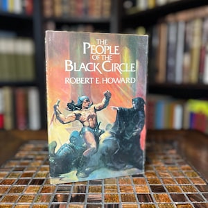 Könnte beinhalten: Ein Taschenbuch mit dem Titel "The People of the Black Circle" von Robert E. Howard. Das Cover zeigt einen muskulösen Mann in einem Lendenschurz, der gegen eine verhüllte Gestalt kämpft.