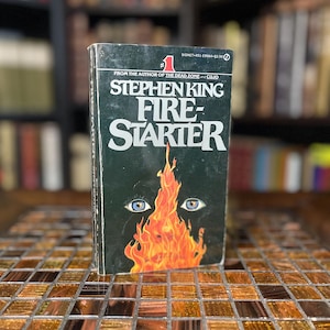 Könnte beinhalten: Ein Taschenbuchcover mit schwarzem Hintergrund und dem Titel "Fire-Starter" in weißer Schrift. Der Autor Stephen King ist unter dem Titel aufgeführt. Das Cover zeigt ein stilisiertes Bild eines Feuers mit zwei Augen, die aus den Flammen schauen.