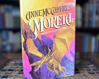 May include: A yellow, orange, and purple book cover with the title "Anne McCaffrey Moreta: Dragonlady of Pern". The cover features a dragon flying in the sky with a rider on its back.