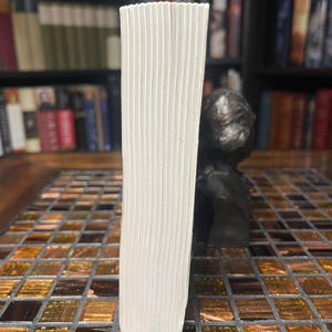 May include: A book with white pages, viewed from the side, showing the edge of the pages. The book is standing upright on a mosaic tile surface. A dark sculpture is in the background, with a bookshelf filled with books.