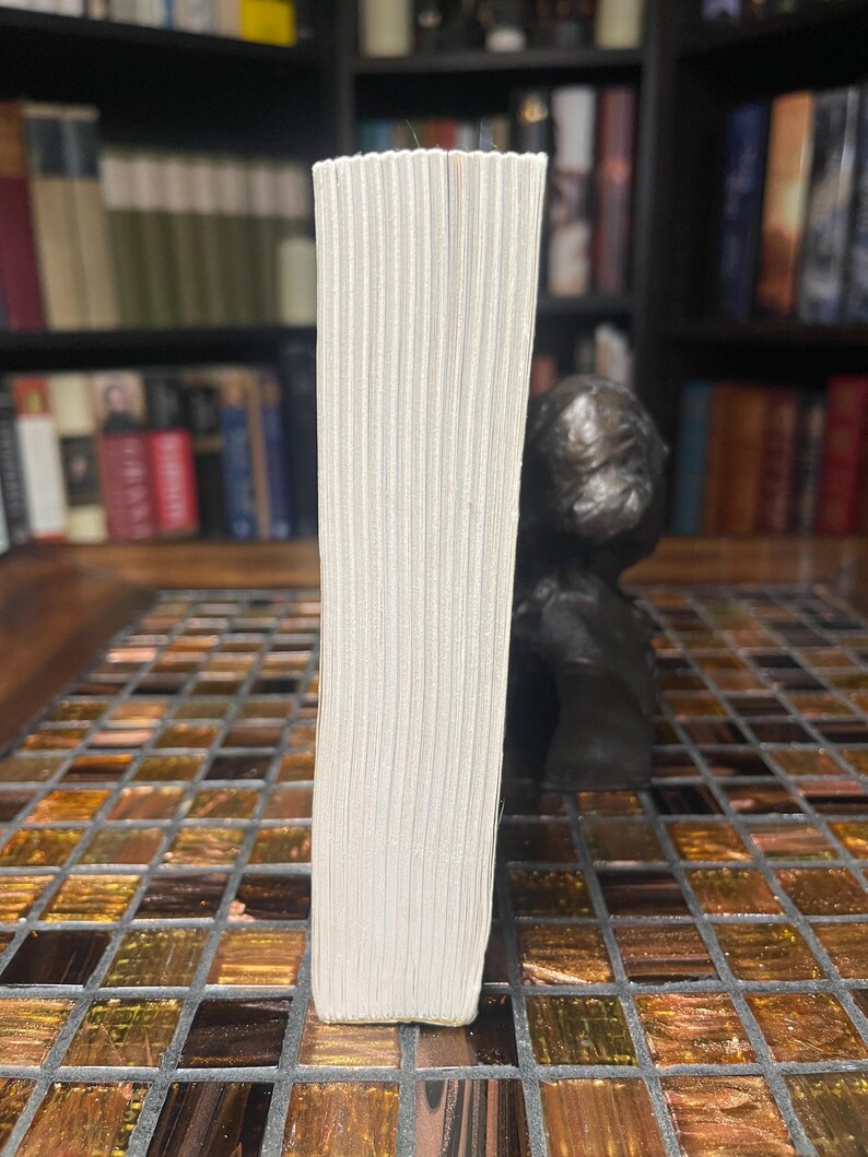 May include: A book with white pages, viewed from the side, showing the edge of the pages. The book is standing upright on a mosaic tile surface. A dark sculpture is in the background, with a bookshelf filled with books.