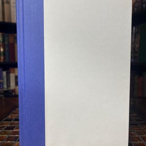 Może przedstawiać: Książka w twardej oprawie z niebieskim grzbietem i białą okładką. Książka stoi pionowo, z grzbietem po lewej stronie. Tło to rozmyty widok p&oacute;łki z książkami.