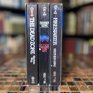 Puede incluir: Tres libros de bolsillo vintage de Stephen King. Los títulos son "The Dead Zone", "Salem's Lot" y "Firestarter". Las lomos negros presentan texto blanco y azul. Los libros se exhiben sobre una superficie de mosaico.
