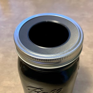 Könnte beinhalten: Ein schwarzes und silbernes Einmachglas mit einem silbernen Deckel. Der Deckel hat eine große Öffnung mit einem schwarzen Gummiring am Rand. Das Glas ist mit "Ball" und "Wide Mouth" beschriftet.