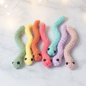Könnte beinhalten: Ein Set aus sechs gehäkelten Regenbogen-Wurm-Spielzeugen mit schwarzen Augen. Die Würmer sind in einem Halbkreis auf einer weißen Marmorfläche angeordnet.