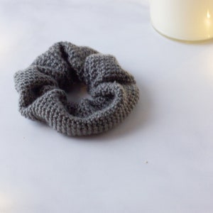 Op de afbeelding: Een grijze gebreide scrunchie, een haaraccessoire, ligt op een wit oppervlak. De scrunchie is gemaakt van een getextureerd, geweven materiaal. Een brandende kaars in een glazen pot staat op de achtergrond.