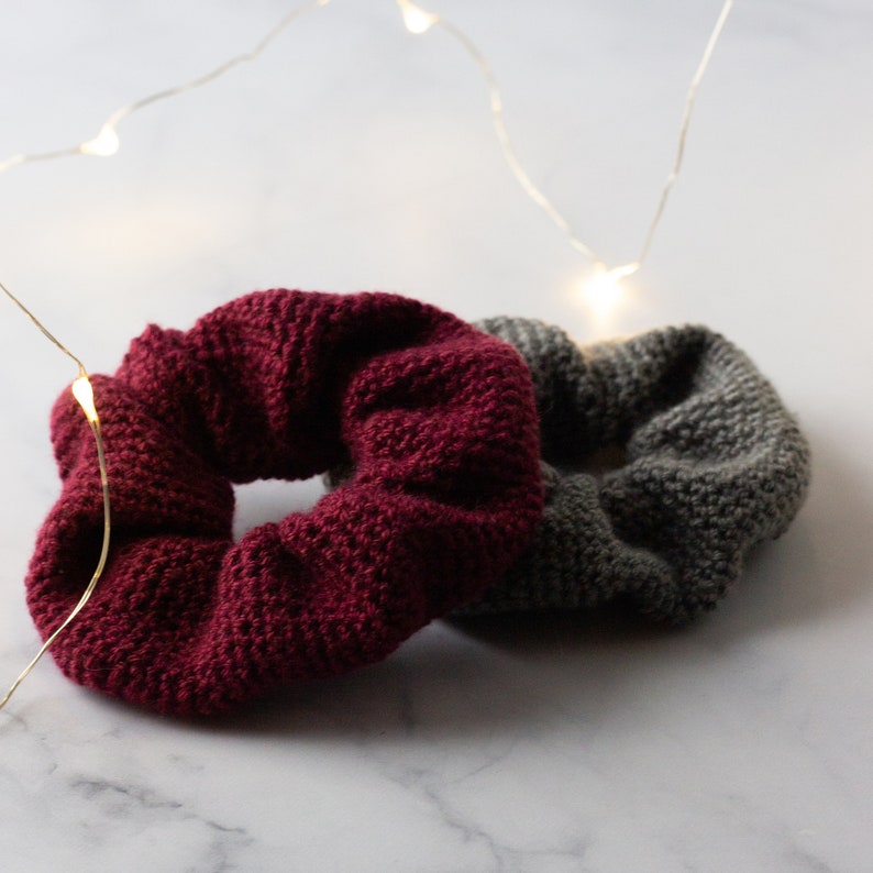 Op de afbeelding: Twee gebreide scrunchies. E&eacute;n is diep bordeauxrood, de andere is middengrijs. De scrunchies zijn gemaakt van dik, getextureerd garen. Het zijn waarschijnlijk haaraccessoires.