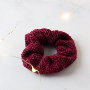Op de afbeelding: Een bordeauxrode, gebreide scrunchie. Het haaraccessoire is gemaakt van een textuurrijk, geweven materiaal. De scrunchie is cirkelvormig en ligt op een wit oppervlak. Slingers met lichtjes zijn op de achtergrond.