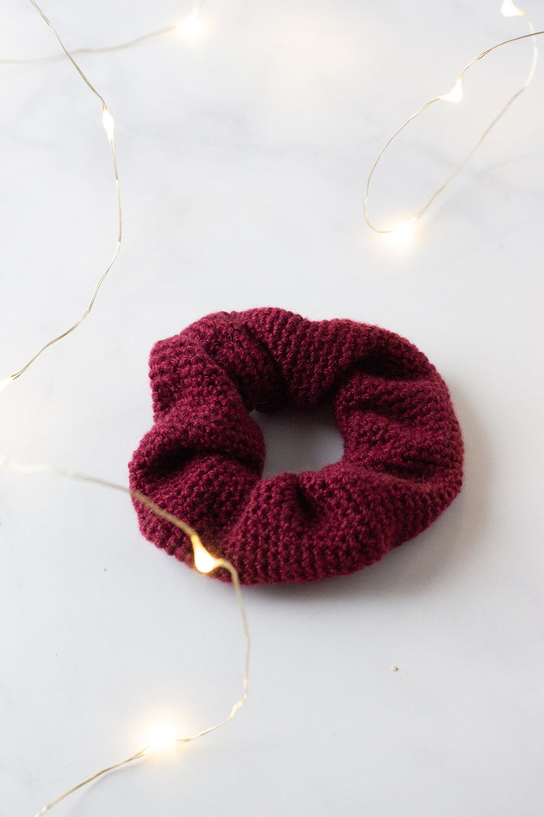 Op de afbeelding: Een bordeauxrode, gebreide scrunchie. Het haaraccessoire is gemaakt van een textuurrijk, geweven materiaal. De scrunchie is cirkelvormig en ligt op een wit oppervlak. Slingers met lichtjes zijn op de achtergrond.