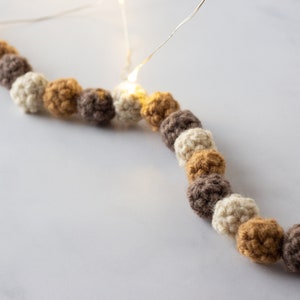 Puede incluir: Una guirnalda de bolas de crochet en beige, marr&oacute;n y amarillo mostaza. La guirnalda est&aacute; enhebrada con luces blancas.