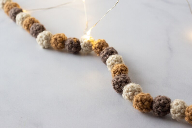 Puede incluir: Una guirnalda de bolas de crochet en beige, marr&oacute;n y amarillo mostaza. La guirnalda est&aacute; enhebrada con luces blancas.