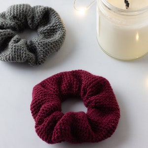 Op de afbeelding: Twee gebreide scrunchies in grijs en bordeaux, naast een kaars in een glazen pot. De scrunchies zijn gemaakt van dik, getextureerd garen. De kaars heeft witte was en een aangestoken lont.