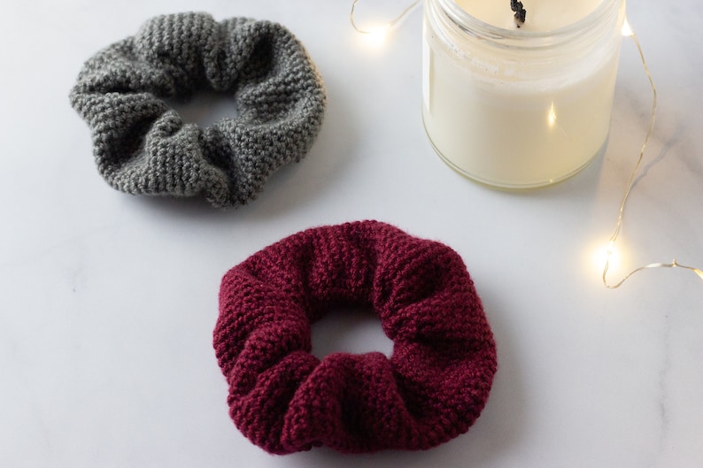 Op de afbeelding: Twee gebreide scrunchies in grijs en bordeaux, naast een kaars in een glazen pot. De scrunchies zijn gemaakt van dik, getextureerd garen. De kaars heeft witte was en een aangestoken lont.