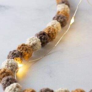 Puede incluir: Una guirnalda de bolas de crochet en color beige, marr&oacute;n y crema est&aacute; enhebrada con una cadena de luces de hadas. Las luces est&aacute;n encendidas y brillan.