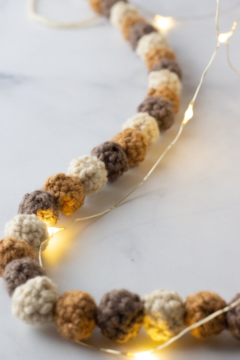Puede incluir: Una guirnalda de bolas de crochet en color beige, marr&oacute;n y crema est&aacute; enhebrada con una cadena de luces de hadas. Las luces est&aacute;n encendidas y brillan.