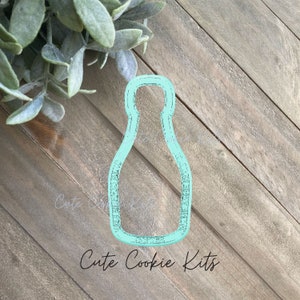 Puede incluir: Un cortador de galletas de plástico azul claro con forma de botella. El cortador de galletas está sobre una superficie de madera.