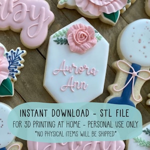 May include: A set of six pink and blue baby shower cookies decorated with flowers, a rattle, and the name "Aurora Ann". The cookies are for 3D printing at home, for personal use only. No physical items will be shipped.