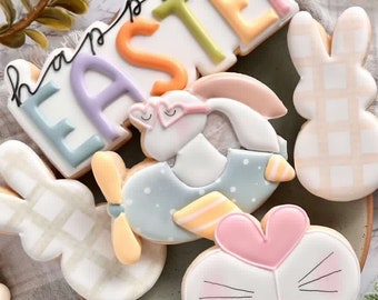 May include: A set of Easter-themed sugar cookies decorated with pastel colors and various designs. The cookies include a bunny wearing glasses, a bunny with a heart-shaped nose, a bunny with a checkered pattern, and a cookie that says "Happy Easter" in rainbow letters.