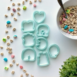 May include: A set of eight light blue plastic cookie cutters in various shapes, including a heart, a horseshoe, a bear, a paw print, a mushroom, a gingerbread man, a crescent moon, and a bell. The cutters are arranged on a white surface.