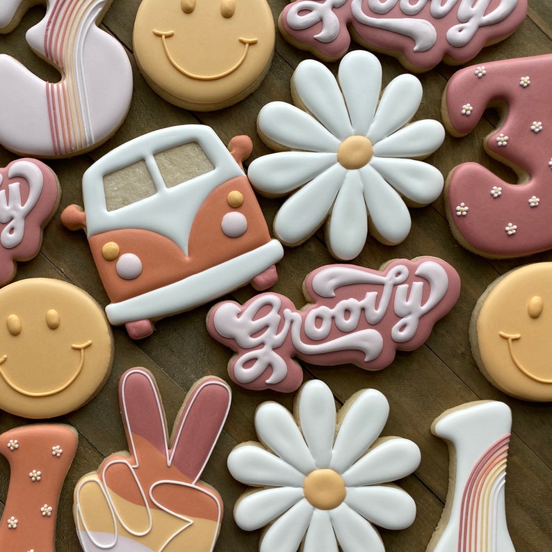 May include: A set of 10 decorated sugar cookies with a groovy theme. The cookies feature a peace sign, a smiley face, a daisy, a vintage van, and the word "groovy" in a retro font. The cookies are decorated in shades of pink, orange, and white.