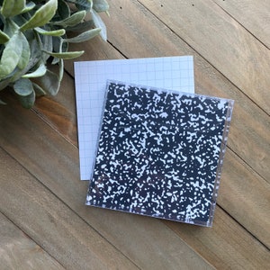 May include: A black and white composition notebook with a grid pattern on the cover. The notebook is on a wooden surface.