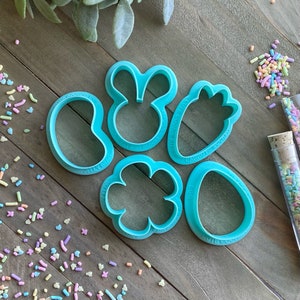 May include: A set of six turquoise blue cookie cutters shaped like a jelly bean, bunny, carrot, flower, and two eggs. The cutters are on a wooden surface with colorful sprinkles scattered around them.