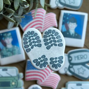 May include: A white cookie decorated with a pair of gray boot prints. The cookie is surrounded by other cookies, including a cookie with an American flag design, a cookie with a soldier's photo, and a cookie with the word "HERO" on it.
