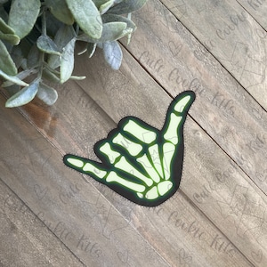 May include: A green glow-in-the-dark skeleton hand cookie cutter in the shape of a shaka sign. The cookie cutter is on a wooden surface.