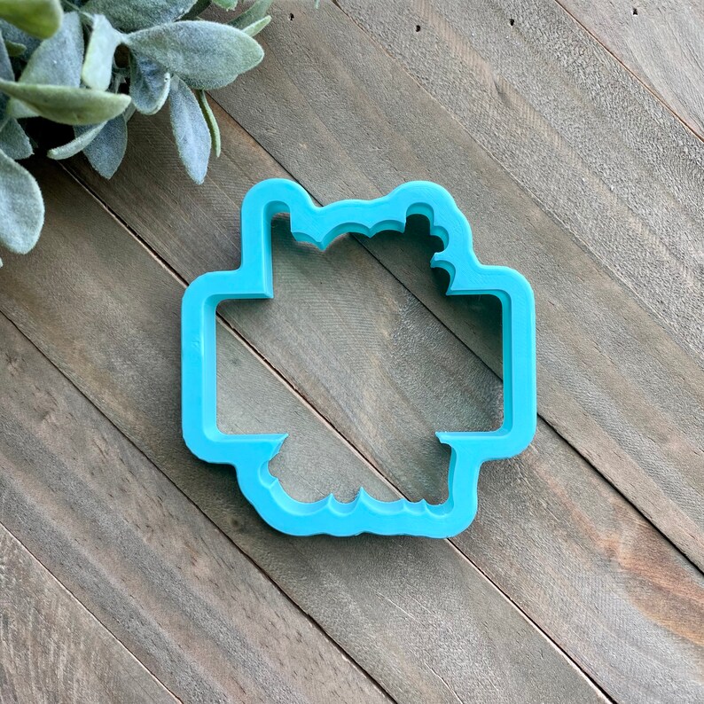 May include: A light blue plastic cookie cutter shaped like a wreath with a rectangular space in the center.