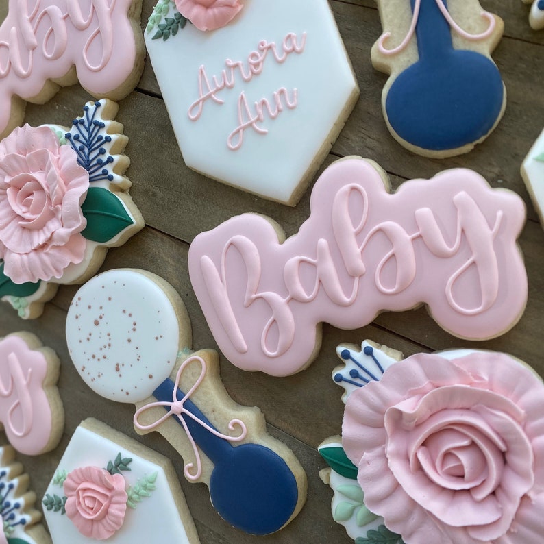 May include: A set of pink and blue baby shower cookies decorated with flowers, a rattle, and the words "Baby" and "Aurora Ann".