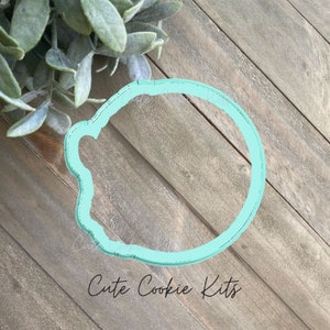 May include: A light blue plastic cookie cutter shaped like a cloud. The cutter is on a wooden surface. "Cute Cookie Kits" is written in black text below the cutter.