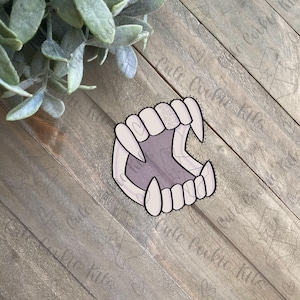 May include: A white and purple illustration of a set of fangs. The fangs are drawn in a cartoon style with sharp points and a slightly open mouth.